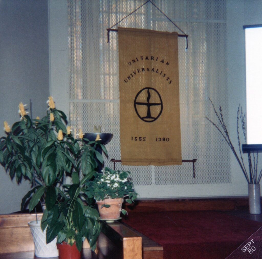 A banner hangs at the front of the church sanctuary on the screen in front of the stained glass windows.  It reads "Unitarian Universalists 1555-1980" and features the flaming chalice.  There are plants on the left and a projector screen on the right.