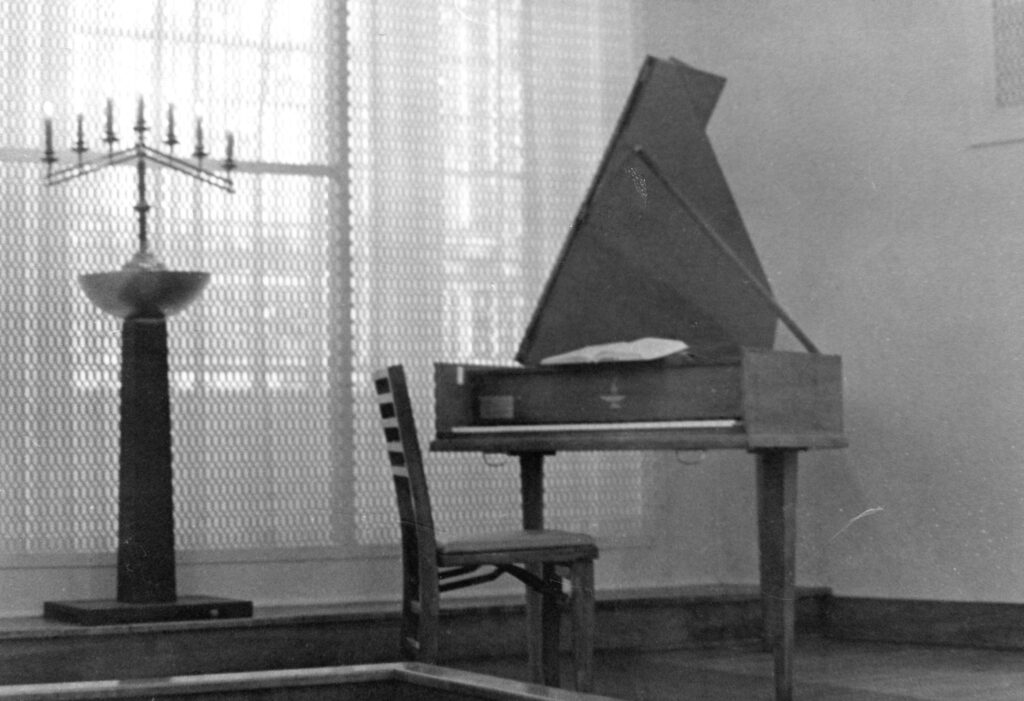 A black and white photograph of a piano and chair at the front of the sanctuary. The stained glass windows are shielded from view by a sun screen.