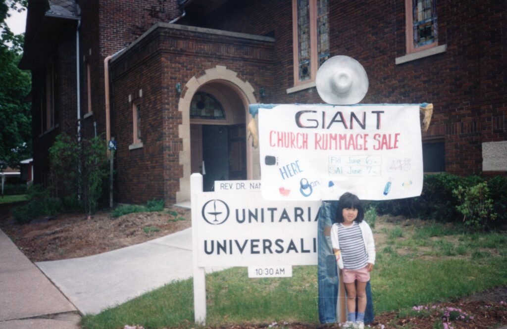 A young girl stands in front of both the Unitarian Universalist Fellowship sign and a large sign advertising a giant church rummage sale.