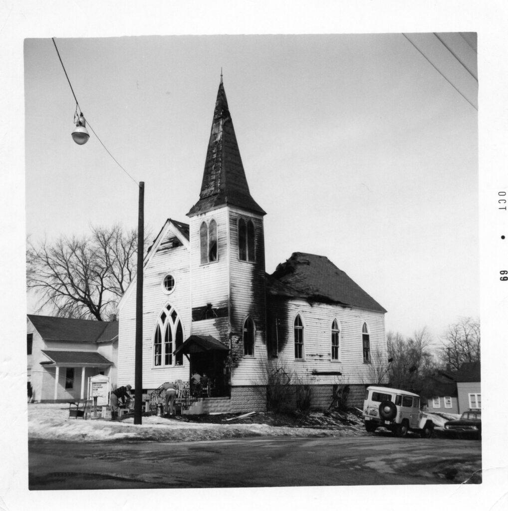A black and white photograph of a classically styled white wooden church that has been badly burned.