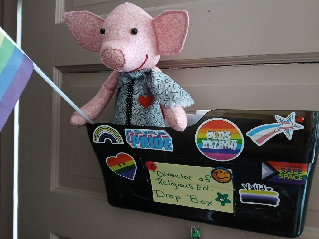 A handmade plush pig in a gray outfit holding a rainbow flag in a door mounted mailbox covered in LGBTQIA+ pride stickers and labelled "Director of Religious Ed. Drop Box."