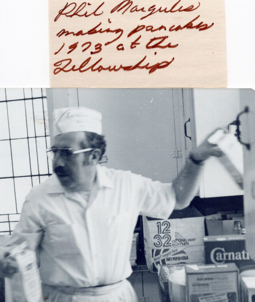 A man in a white shirt and ice-cream deliveryperson hat cooks pancakes in the church kitchen.