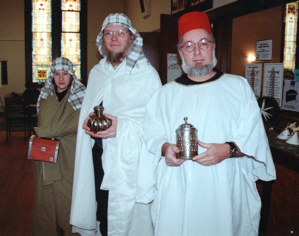Three men in "Wise Man" costumes of white robes, two with plaid headscarves and one in a fez, carry containers during the Christmas pageant.